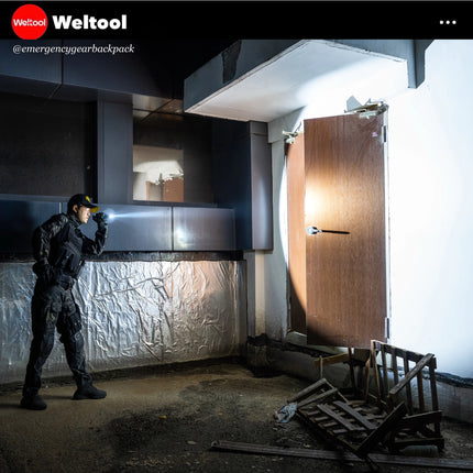 Person in tactical gear standing in a dimly lit room with a large open door.